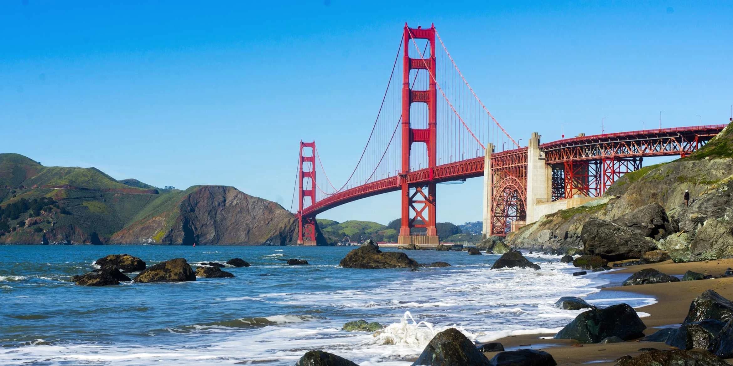 The completed Golden Gate Bridge spanning blue water, seen from a rocky beach beneath a clear sky.