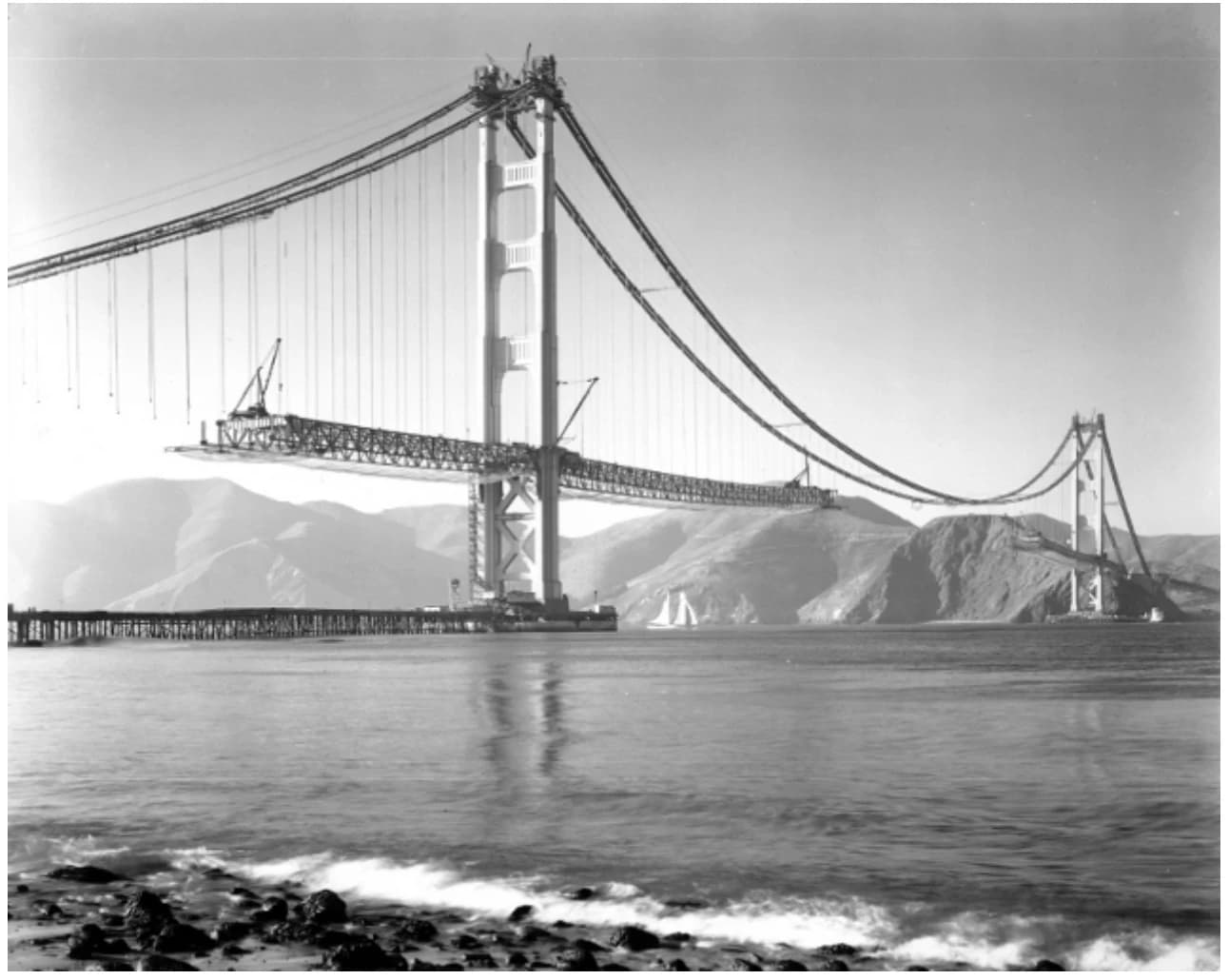 The Golden Gate Bridge under construction, with its two towers and cables in place and the roadway extending partway across the water.